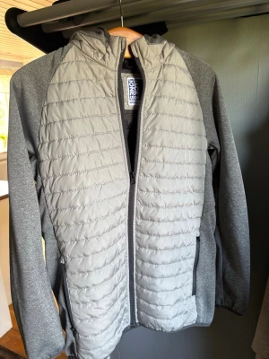 Grå cardigan - Snygg grå cardigan från Jack & Jones med quiltad front och huva. Jackan har dragkedja framtill, två sidofickor och är tillverkad i en mix av slätt och melerat material. Ända defekten är hängaren vilket syns på bilden men annars i väldigt bra skick. Hör av er vid frågor!