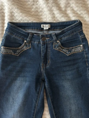Blå jeans med broderade fickor Madlady - Snygga blå jeans från Madlady med unika broderade detaljer på både fram- och bakfickor. Bakfickorna har coola broderade vingar i vitt och silver, och framfickorna har mönstrad kant. Klassisk femficksmodell i stretchigt denimtyg. Inte fel på dem