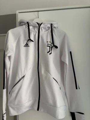 Vit hoodie med Juventus-tryck - Snygg vit hoodie med svart Juventus-tryck och två stjärnor på bröstet. Dragkedja framtill, svarta detaljer och snören i huvan. Två fickor med dragkedja och Adidas-logga på bröstet. Perfekt för träning eller chill. Köpte den på en affär i Spanien 
