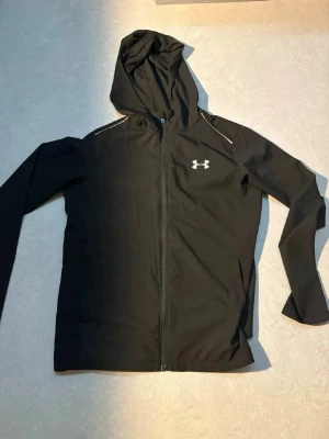 Svart vindjacka från Under Armour - Snygg svart vindjacka från Under Armour med huva och hel dragkedja framtill. Jackan har vita detaljer och logga på bröstet samt stort UNDER ARMOUR-tryck på ryggen. Tillverkad i lätt och smidigt material, perfekt för blåsiga dagar.