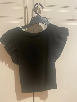 Svart ribbad topp från Zara - Säljer en svart ribbad topp från Zara med volangdetaljer på ärmarna. Toppen har en rund halsringning och är tillverkad i ett mjukt, stretchigt material. Perfekt för dig som gillar stilrena plagg med en twist.