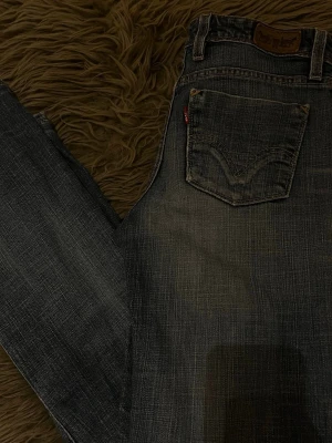 Blå Levi's raka jeans - Säljer ett par klassiska blå Levi's jeans med raka ben och tydliga slitningar för en vintage look. Dem är ett par jeans köpta under 90-talet early 2000’s. Jeansen har fem fickor, kontrastsömmar och den ikoniska röda Levi's-lappen på bakfickan. Tillverkade i slitstark denim, perfekta för någon som gillar vintage kläder.