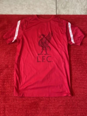 Röd Liverpool FC t-shirt - Säljer en röd t-shirt med Liverpool FC-logga på bröstet och texten L.F.C. Tryckta vita detaljer på ärmarna. Perfekt för dig som älskar fotboll och vill visa ditt stöd för laget. Tillverkad i ett lätt och bekvämt material som passar till träning eller match.