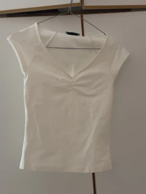 Vit v-ringad topp från Bershka - Snygg vit topp från Bershka med v-ringning och lätt rynkdetalj vid bysten. Modellen är ärmlös och har en slim passform som framhäver figuren. Perfekt att matcha med jeans eller kjol för en clean och fräsch look.