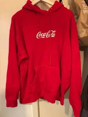 Röd Coca-Cola hoodie - Finns en LITEN svart prick! Syns på bild2 .      Snygg röd hoodie med klassisk Coca-Cola-logga i vitt på bröstet. Hoodien har huva med dragsko och en stor magficka framtill. Tillverkad i mjukt material som känns skönt mot huden. Perfekt för dig som gillar streetwear och vill sticka ut. 