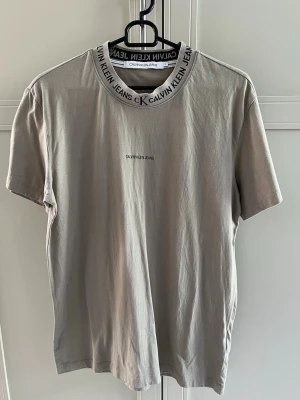 Beige t-shirt från Calvin Klein Jeans - Snygg beige t-shirt från Calvin Klein Jeans med logga på bröstet och tryckt text runt kragen. Klassisk passform och korta ärmar, tillverkad i mjuk bomull. 