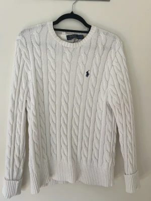 Vit kabelstickad tröja Polo Ralph Lauren - Kabelstickad vit tröja från Polo Ralph Lauren med rund halsringning och marinblå broderad logga på bröstet. Tröjan har långa ärmar, ribbade muddar och nederkant. Klassisk och tidlös design i mjukt material.