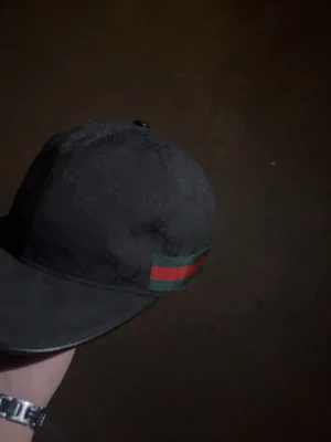 Svart keps med Gucci-mönster - Snygg svart keps från Gucci med det klassiska GG-mönstret över hela kepsen och en grön-röd rand på sidan. Justerbar rem baktill för perfekt passform. Materialet känns slitstarkt och kepsen har en böjd skärm.