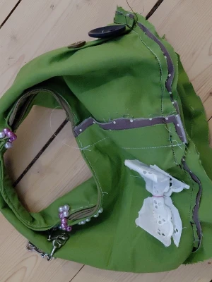 Grön axelväska med spetsrosett - Unik grön handväska i tyg med dekorativa sömmar och en vit spetsrosett framtill. Väskan har dragkedja, pärl- och kedjedetaljer på handtaget samt en knappstängning. Perfekt för dig som gillar personliga accessoarer med DIY-känsla.