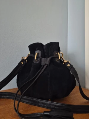 Svart mocka bucket bag med axelrem - Snygg svart bucket bag i mjuk mocka med detaljer i läder och guldfärgade metalldelar. Väskan har justerbar axelrem och dragskostängning. Rund botten med dekorativa nitar och läderband. Perfekt för dig som gillar en unik och trendig look.