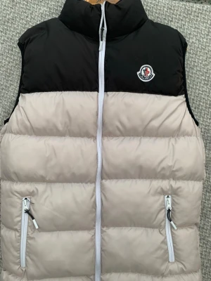 Beige och svart dunväst från Moncler - Snygg dunväst från Moncler i beige och svart med hög krage och vita dragkedjor. Västen har två fickor framtill och Moncler-logga på bröstet. Perfekt för lager på lager och ger en clean look till din outfit.
