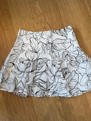 Vit blommig kortkjol i linneblandning - Säljer en vit kortkjol med svart blommigt mönster från H&M. Kjolen är i linneblandning och har en lätt utsvängd form som ger en skön och luftig känsla. Perfekt för varma dagar och enkel att matcha med olika toppar.
