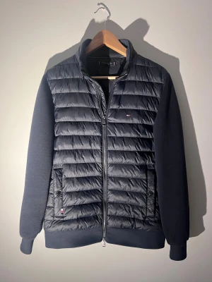 Blå Cardigan Tommy Hilfiger - Blå pufferjacka från Tommy Hilfiger i storlek S. Jackan har quiltad framsida med dragkedja, ribbade muddar och diskret logga på bröstet. Perfekt för höst och vinter. Mycket liv kvar att ge | passar ca 172-182 | Har tecken på användning | nopprig på rygg, och armarna | Nypriss : 2749