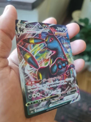Pokémonkort Umbreon VMAX japansk - Säljer ett japanskt Pokémonkort med Umbreon VMAX-motiv. Kortet har en färgstark och glansig design med Umbreon i svart, blått och gult mot en regnbågsskimrande bakgrund. Perfekt för samlare eller Pokémonfans.