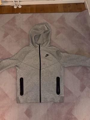 Grå Nike hoodie med dragkedja - Snygg grå hoodie från Nike med hel dragkedja framtill och två svarta dragkedjeförsedda fickor. Klassisk Nike-logga på bröstet och huva för extra komfort. Tillverkad i mjukt material, perfekt för en avslappnad stil.