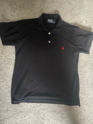 Svart pikétröja från Polo Ralph Lauren - Klassisk svart pikétröja från Polo Ralph Lauren med vit knappslå och den ikoniska röda logon broderad på bröstet. Tröjan har korta ärmar och mjukt bomullsmaterial som känns skönt mot huden. Perfekt till jeans eller shorts.