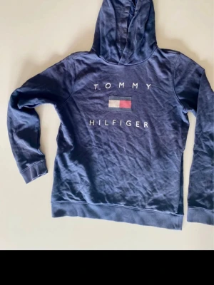Blå hoodie Tommy Hilfiger 16 års 176 - Blå hoodie från Tommy Hilfiger med klassisk logga och text på bröstet. Tröjan har huva, långa ärmar och ribbade muddar. Tillverkad i mjukt bomullsmaterial, perfekt för träning eller chill.