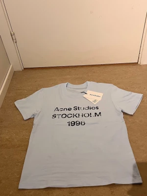 Ljusblå Acne Studios t-shirt - Säljer en ljusblå t-shirt från Acne Studios med tryck i svart text på bröstet där det står 'Acne Studios STOCKHOLM 1996'. Klassisk rund hals och korta ärmar. Perfekt för dig som gillar stilrena plagg med en twist.