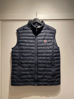 Mörkblå Colmar väst - Snygg mörkblå dunväst med quiltat mönster och hög krage. Västen har två dragkedjeförsedda fickor framtill och en liten logotyp på bröstet. Perfekt att slänga över en hoodie eller tröja för extra värme.