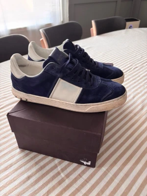 Valentino flycrews - Klassiska och tidlösa Valentino Flycrew sneakers i mörkblå mocka med den ikoniska vita skinnranden över sidan och Rockstud-detaljer längst bak på hälen. En mycket eftertraktad modell. Skorna är användna och säljs i befintligt skick. Det finns normalt slitage på mockan, innersulan och undersulan.  Obs: En av de små nitarne (Rockstuds) på baksidan av hälen har lossnat. Detta går dock enkelt att ersätta genom att köpa en ny nit online eller hos en skomakare.
