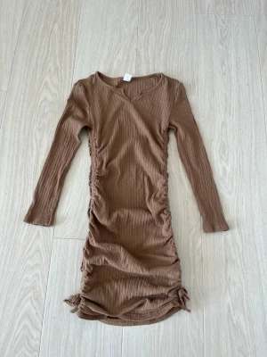 Ribbad brun klänning med snörning - Snygg ribbad klänning i brun färg med långa ärmar och snörningsdetaljer på sidorna. Klänningen har en kroppsnära passform och är tillverkad i ett mjukt, stretchigt material som ger en bekväm känsla.