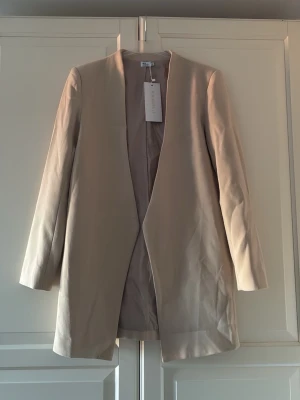 Öppen kavaj från Chiquelle - Stilren öppen kavaj från Chiquelle med rak passform och öppen front utan knappar. Kavajen har långa ärmar och är tillverkad i ett mjukt, slätt tyg som ger en clean look. Storlek M. Helt ny med etiketten kvar.