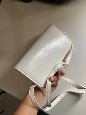 Beige axelväska i konstläder - Stilren beige axelväska i konstläder med enkel, rektangulär form och justerbar axelrem. Väskan har guldfärgade metalldetaljer och en slät yta som ger en modern look. Perfekt för dig som gillar minimalistisk stil. Helt ny luktar lite PU leder men försvinner efter att du har använd det några gånger. Skriv gärna till mig innan köp
