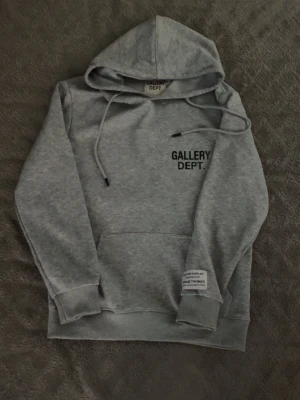 Grå hoodie från Gallery Dept. - Snygg grå hoodie från Gallery Dept. med svart tryck på bröstet och en patch på ärmen. Klassisk känguruficka framtill, justerbar huva med dragsko och ribbade muddar. Perfekt för en avslappnad streetwear-look.