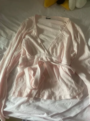 Ljusrosa omlottblus med knytband - Söt ljusrosa omlottblus med knytband i midjan och djup v-ringning. Blusen har långärmade ärmar och är tillverkad i ett lätt, krinklat tyg som ger en avslappnad men trendig look. Perfekt att styla med jeans eller kjol för en fräsch outfit. 