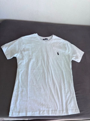 Vit polo t-shirt - Klassisk vit t-shirt i bomull med rund hals och korta ärmar. På bröstet finns en liten broderad logga i svart som ger en stilren touch. Perfekt basplagg till garderoben och enkel att matcha med allt.