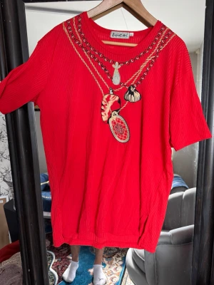 Röd t-shirt med broderade kedjor - Cool röd t-shirt från Wico med broderade kedjor och hängen framtill. T-shirten har korta ärmar och är gjord i ribbad bomull som ger en skön känsla. Perfekt för dig som vill sticka ut med ett unikt motiv och färg.