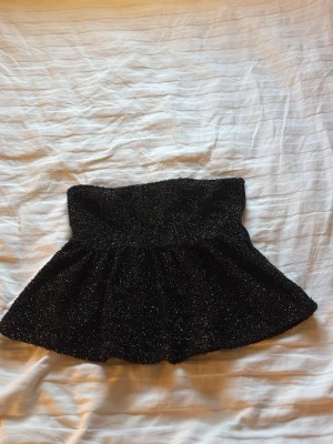 Svart glittrig topp med dragkedja - En svart glittrig topp med lätt utsvängd form. Toppen har en diskret dragkedja i sidan och är fodrad för extra komfort. Perfekt för dig som vill sticka ut med lite glitter.
