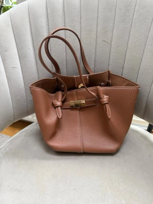 Brun bucket handväska i konstläder - Snygg brun handväska i bucket-modell med guldfärgat spänne och dragkedja. Väskan har två smala axelband och är tillverkad i konstläder med struktur. Invändigt finns ett fack med dragkedja och magnetknäppning. Perfekt för dig som gillar stilrena accessoarer. Populär väska från Lindex. Kommer utan den långa bandet. 