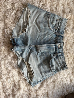 Ljusblåa  jeansshorts - Säljer ett par ljusblåa jeansshorts med hög midja och råa, slitna kanter. Shortsen har klassisk femficksdesign och fransiga detaljer vid bensluten för en avslappnad look. Perfekta för varma dagar och festivaler.