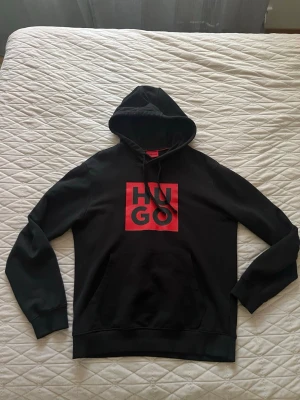 Svart hoodie från HUGO med röd logga - Snygg svart hoodie från HUGO med stor röd logga på bröstet. Klassisk huva med dragsnören och en stor magficka framtill. Tillverkad i mjukt material som känns skönt mot huden. Perfekt för dig som gillar streetwear och vill sticka ut.