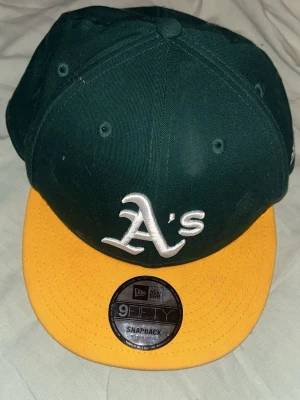 Grön och gul snapback keps New Era - Snygg snapback keps från New Era med mörkgrön krona och gul skärm. Framtill sitter ett broderat vitt 'A's'-emblem och på sidan finns New Era-loggan. Klassisk 9FIFTY-modell med justerbar baksida. Perfekt för dig som gillar sportig stil.