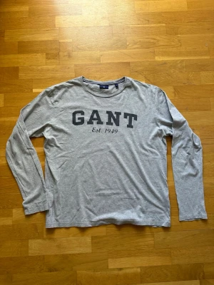 Grå långärmad t-shirt från GANT - Snygg grå långärmad t-shirt från GANT med tryck 'GANT Est. 1949' på bröstet.    Storlek: XL Skick: Mycket bra, inga hål eller fläckar Passform: Normal  Har du några frågor eller vill ha fler bilder? Tveka inte på att höra av dig!