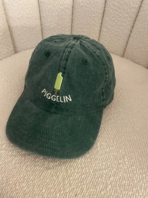 Grön keps med Piggelin-brodyr - Säljer en grön keps med broderad Piggelin-glass och texten 'PIGGELIN' framtill. Kepsen har böjd skärm, justerbart spänne bak och är tillverkad i mjuk bomull. 
