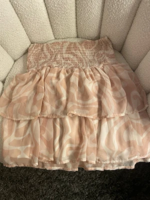 Beige volangkjol från KappAhl - Söt volangkjol från KappAhl i rosa och vitt med abstrakt mönster. Kjolen har bred smock i midjan och dubbla lager av tunn, luftig chiffong som ger en flowy känsla. Perfekt för dig som gillar en feminin och trendig stil.