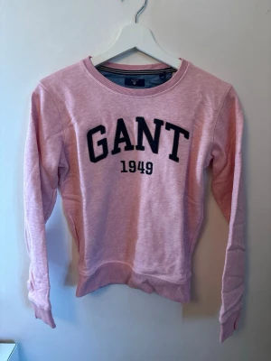 Rosa sweatshirt från GANT - Säljer en ljusrosa sweatshirt från GANT med svart broderad logga och årtalet 1949 på bröstet. Tröjan har rund halsringning, långa ärmar och ribbade muddar vid ärmslut och nederkant. Perfekt för en avslappnad och stilren look.
