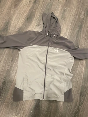 Grå vindjacka från Under Armour - Snygg tvåfärgad vindjacka från Under Armour i ljusgrått och mörkgrått. Jackan har huva med dragsko, hel dragkedja framtill och två sidofickor. Under Armour-logga på bröstet och elastiska muddar vid ärmsluten. Perfekt för blåsiga dagar.