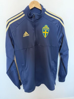 Blå träningströja Sverige Adidas M - Säljer en blå träningströja från Adidas med Sveriges landslagsemblem på bröstet. Tröjan har halv dragkedja, gula detaljer på axlarna och Adidas-logga. Perfekt för träning eller att bära till matchen. Tillverkad i funktionsmaterial.