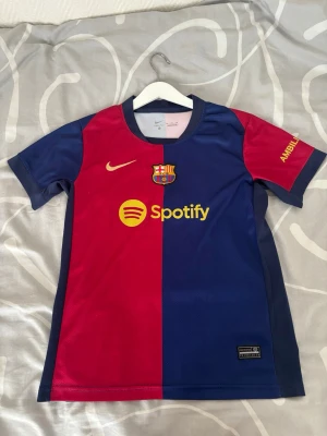 FC Barcelona matchtröja Lamine Yamal - Säljer en  FC Barcelona tröja från Nike med Lamine Yamal och nummer 19 på ryggen. Tröjan är röd och blå med gula detaljer, korta ärmar och klubbmärke samt sponsorloggor på bröstet. Perfekt för dig som älskar fotboll och Barca! Använd några gånger, och har en mindre defekt på baksidan.