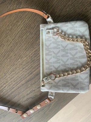 Michael Kors axelväska  - Snygg MK väska med guldig kedja framtill. Väskan har en dragkedja upptill och justerbar axelrem i brunt läder med nitar. Tappat lite färg och därav priset 