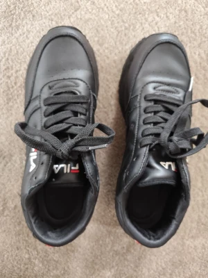 Svarta Fila sneakers i läder - Snygga svarta sneakers från Fila i läder med klassisk låg profil och svarta skosnören. Diskret Fila-logga i vitt och rött på sidan. Perfekta för dig som gillar en clean och sportig look.
