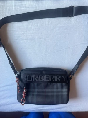 axelväska från Burberry - Snygg svart axelväska från Burberry med klassiskt rutmönster och stor logga framtill. Väskan har justerbar axelrem och detaljer i rep, vilket ger en cool touch. Tillverkad i slitstarkt tygmaterial och perfekt för att bära det viktigaste.