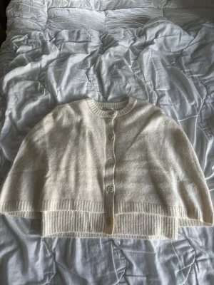 Krämvit cape med knappar - Mysig och oversized krämvit cape med rund halsringning och stora ribbade muddar. Koftan har knäppning framtill med fyra matchande knappar och är tillverkad i ett mjukt, fluffigt material som känns lyxigt. Perfekt för lager på lager och chill dagar.
