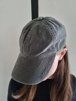 Grå keps från Acne Studios - Snygg grå keps från Acne Studios i tvättad bomull med böjd skärm och broderad logga på sidan. Kepsen har även en liten silverfärgad hjärtberlock med Acne Studios ingraverat, vilket ger en unik detalj. Perfekt accessoar för en avslappnad streetwear-look.