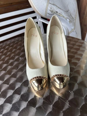 Beige pumps med gulddetalj från NLY - Pumps med spetsig tå och en guldfärgad metallhätta med kedjedetalj. Skorna har en smal klack som är ca 12-13 cm, lätt fläckar förekommer