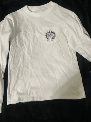 Vit långärmad topp från Chrome Hearts - Vit långärmad topp från Chrome Hearts med stor svart logga på ryggen och mindre logga på bröstfickan. Ärmarna har färgglada tryck i blått, rött och svart. T-shirten är i bomull och har en normal passning. Perfekt för dig som gillar streetwear och coola detaljer.